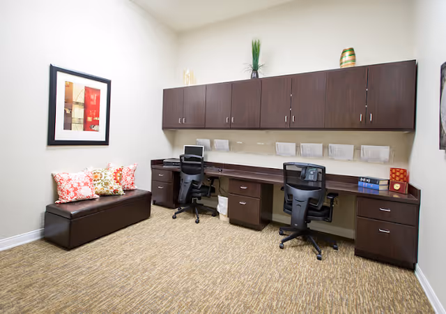 A small office area with two black office chairs at a long dark wood desk with multiple drawers. Above the desk are dark wood cabinets mounted on the wall. On the left side of the room is a brown bench with three decorative pillows in red, white, and green patterns. A framed abstract artwork hangs on the wall above the bench. The floor is carpeted in a beige pattern, and the walls are painted light beige.