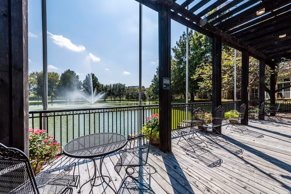 Outdoor covered patio area with black metal tables and chairs overlooking a pond with a water fountain. The patio has wooden flooring and is decorated with potted flowers. Trees and a clear blue sky are visible in the background.