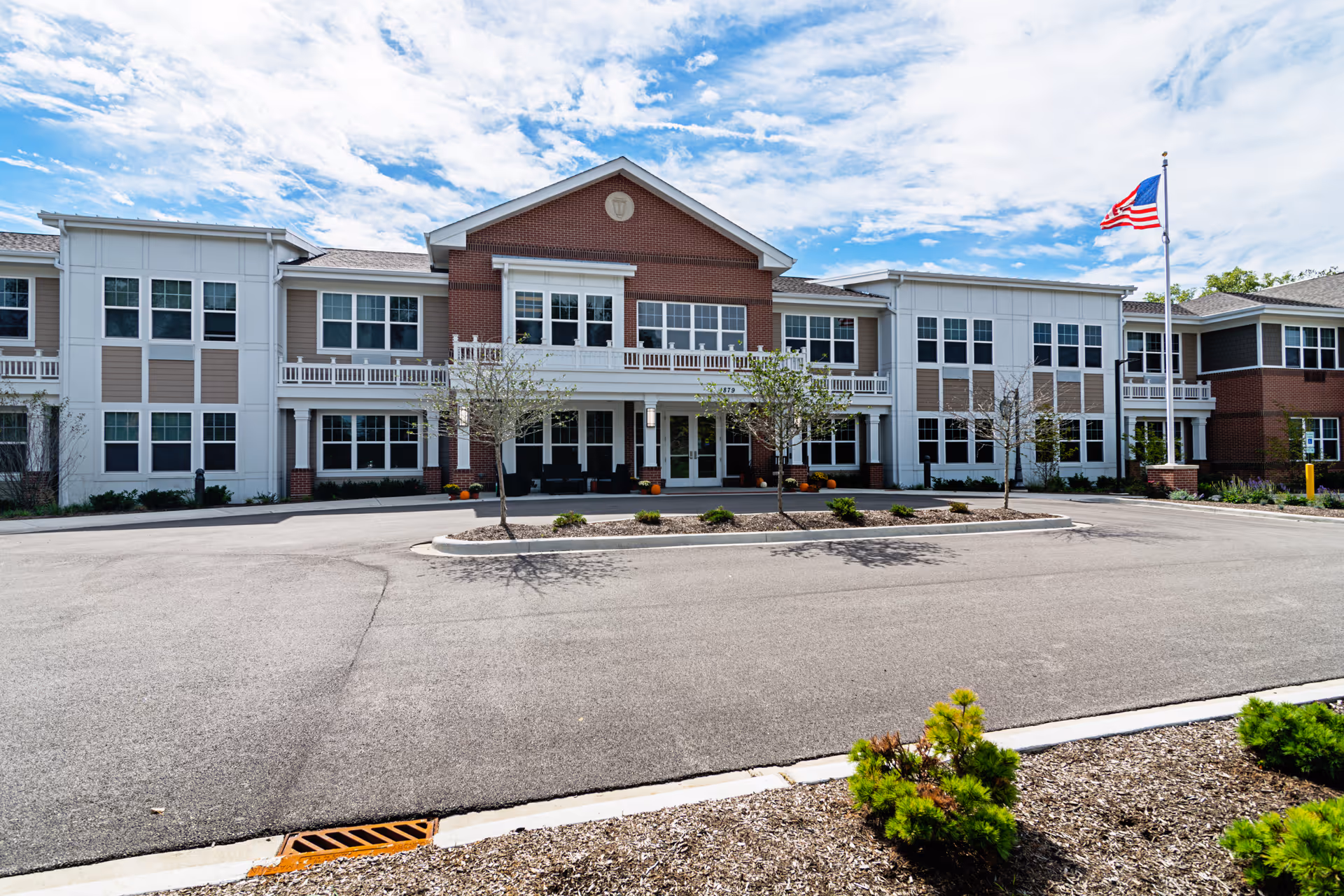 Front exterior view of a two-story senior living facility building with a combination of brick and white siding. The building has multiple windows, a central entrance with a covered porch, small trees and shrubs in front, and an American flag on a flagpole to the right. The sky is partly cloudy.