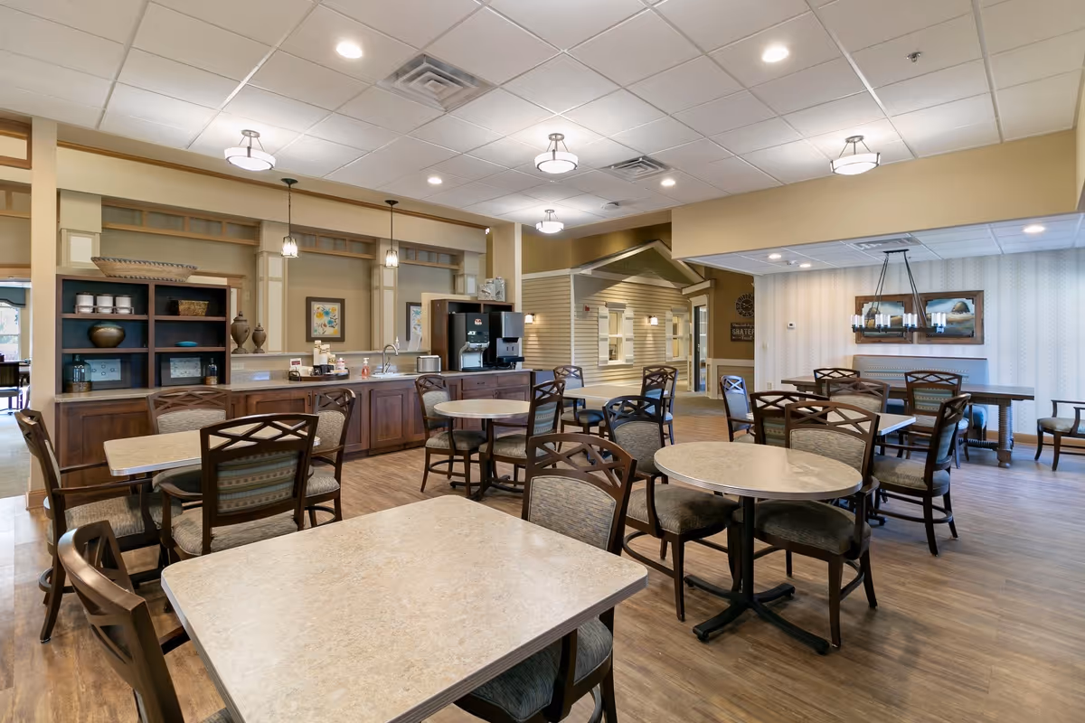 A spacious dining area in St. Andrew’s Village featuring multiple tables and chairs arranged neatly on a wooden floor. The room has a light-colored ceiling with recessed lighting and hanging pendant lights. There is a counter with a coffee machine and decorative items along one wall, and a small house-like structure is visible in the background.