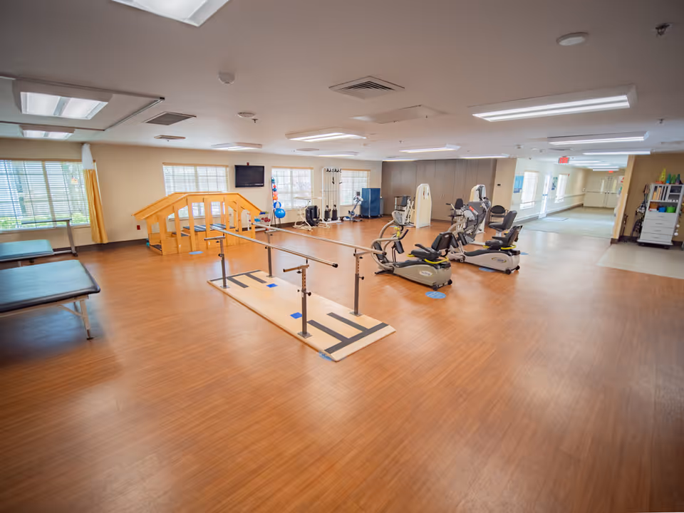 A spacious rehabilitation or physical therapy room with wooden flooring, parallel bars for walking exercises, exercise bikes, therapy tables, and various exercise equipment. The room has large windows with blinds, bright overhead lighting, and a TV mounted on the wall.