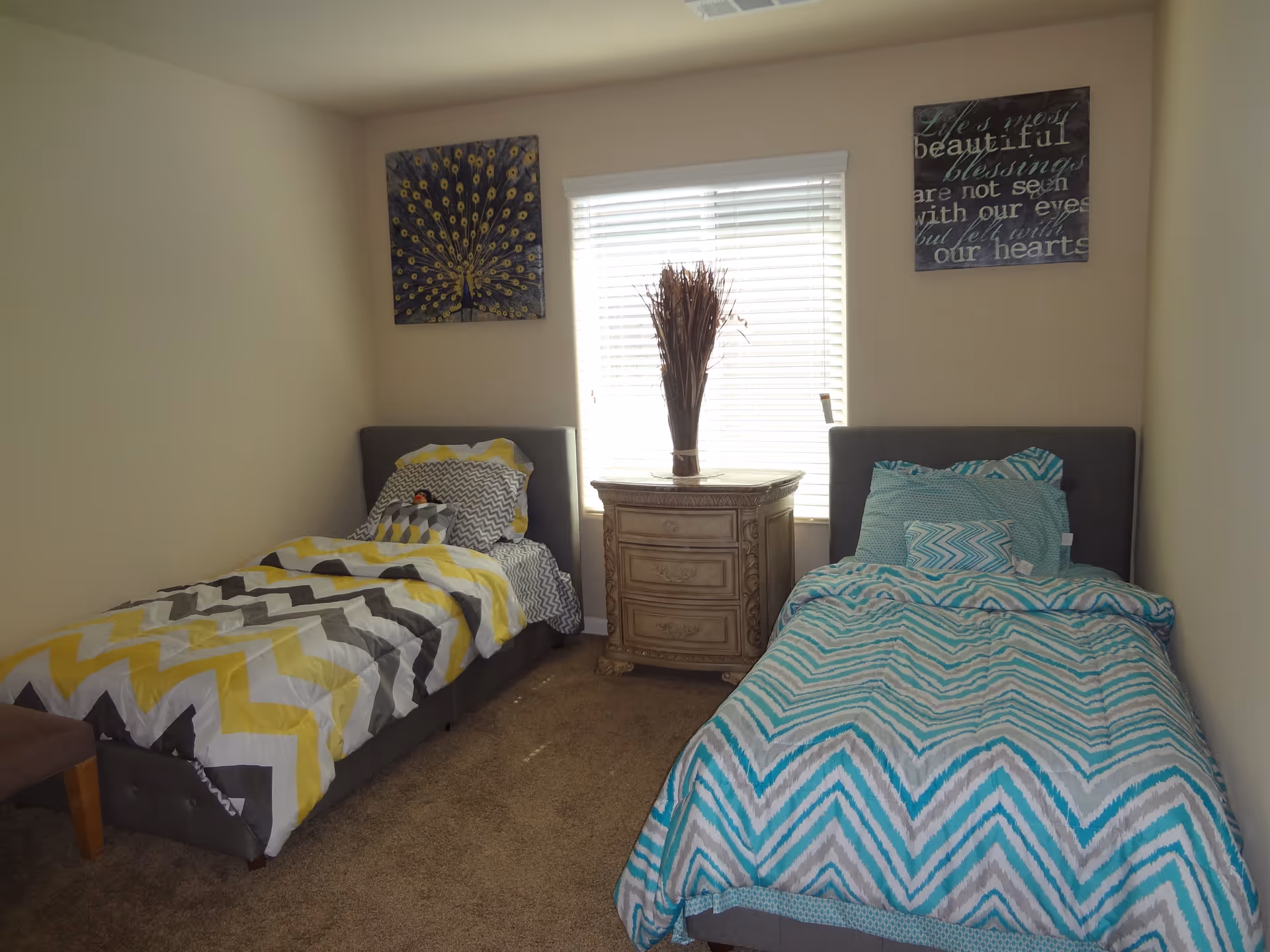 A bright bedroom with two twin beds, a nightstand beneath a window, and framed wall art above each bed.