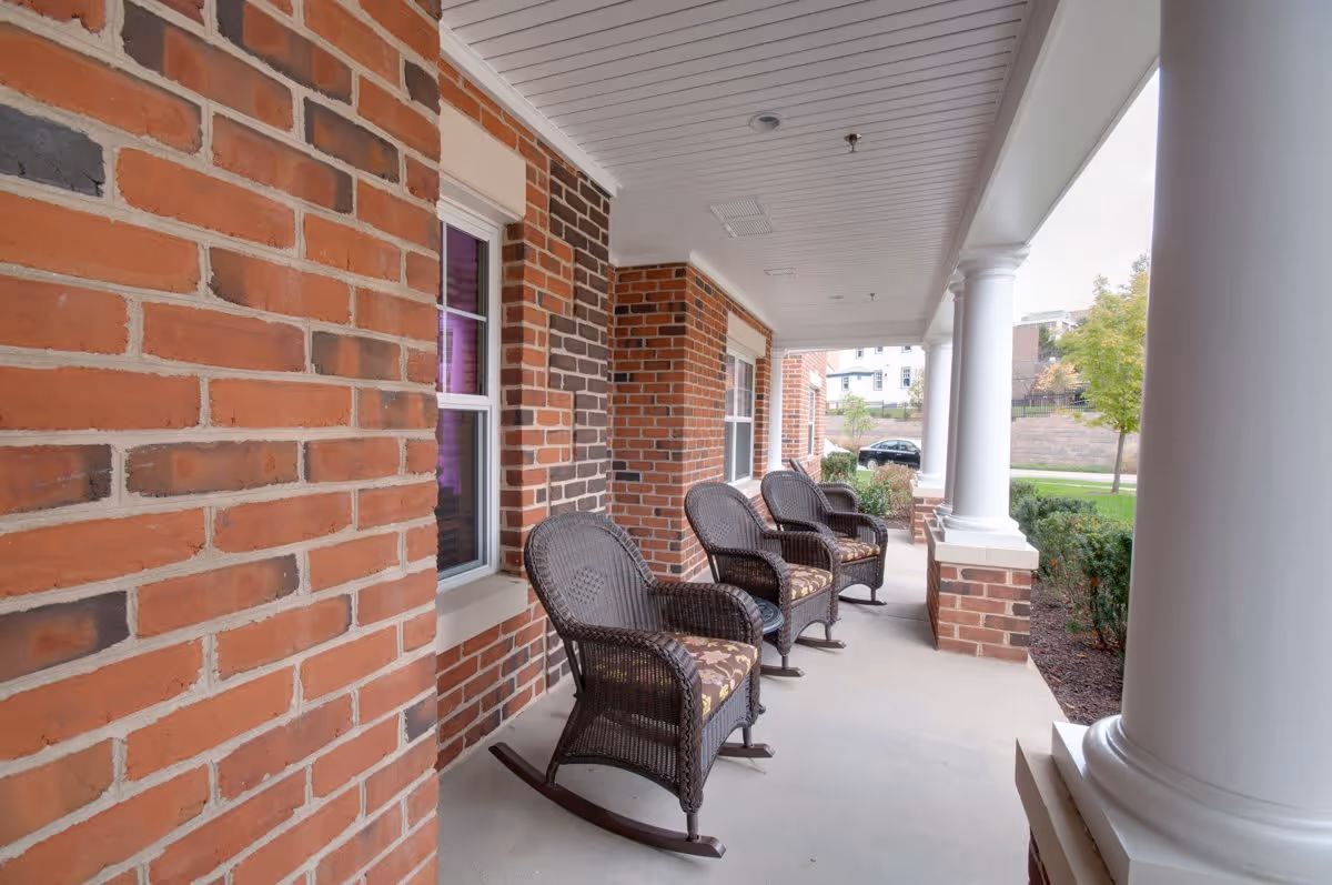 A covered outdoor porch area with four dark wicker rocking chairs with floral cushions lined up against a brick wall. The porch has white columns and overlooks a green lawn with trees and a parked car in the background.