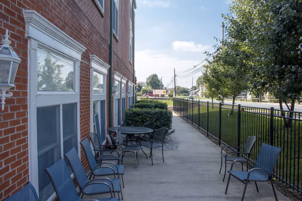 Outdoor patio area alongside a brick building with several metal chairs and a round table. The patio is bordered by a black metal fence and there are bushes and trees along the fence. A sign for Whitehall Manor is visible in the distance near the street.