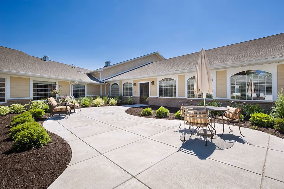 Outdoor patio area at The Village at Hamilton Pointe featuring a concrete walkway, garden beds with green shrubs, and outdoor seating with a table and umbrella under a clear blue sky.