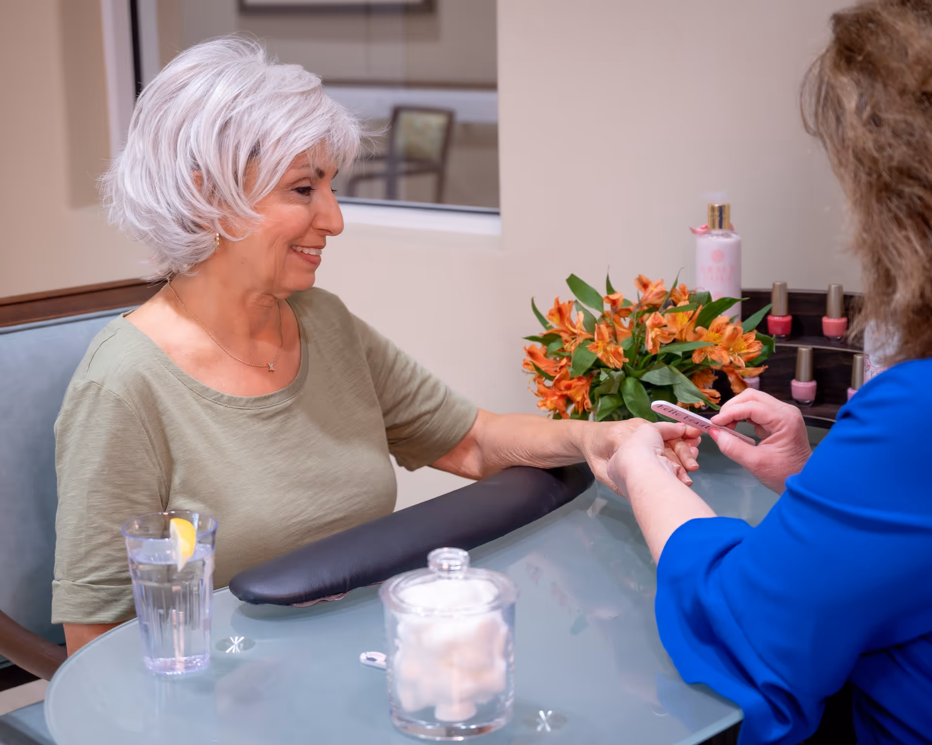 An elderly woman with short white hair is sitting at a glass table receiving a manicure from another person wearing a blue shirt. On the table, there is a glass of water with a lemon slice, a container with cotton balls, a bouquet of orange flowers, and several bottles of nail polish.