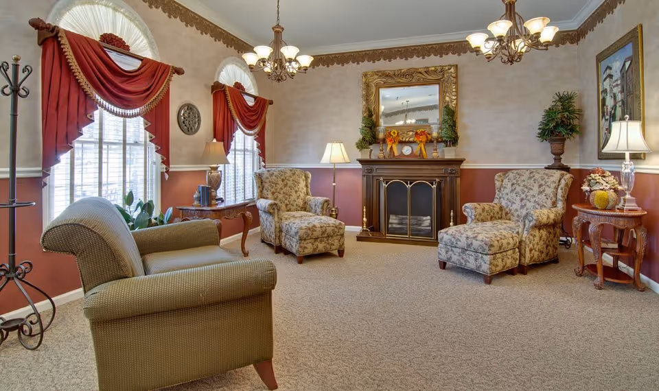 A cozy living room with patterned armchairs and ottomans arranged around a dark wood fireplace with a decorative gold-framed mirror above it. The room features two large windows with red drapes and valances, beige walls with a maroon lower half, carpeted floor, and several lamps providing warm lighting. There are also small tables with decorative items and plants in the corners.