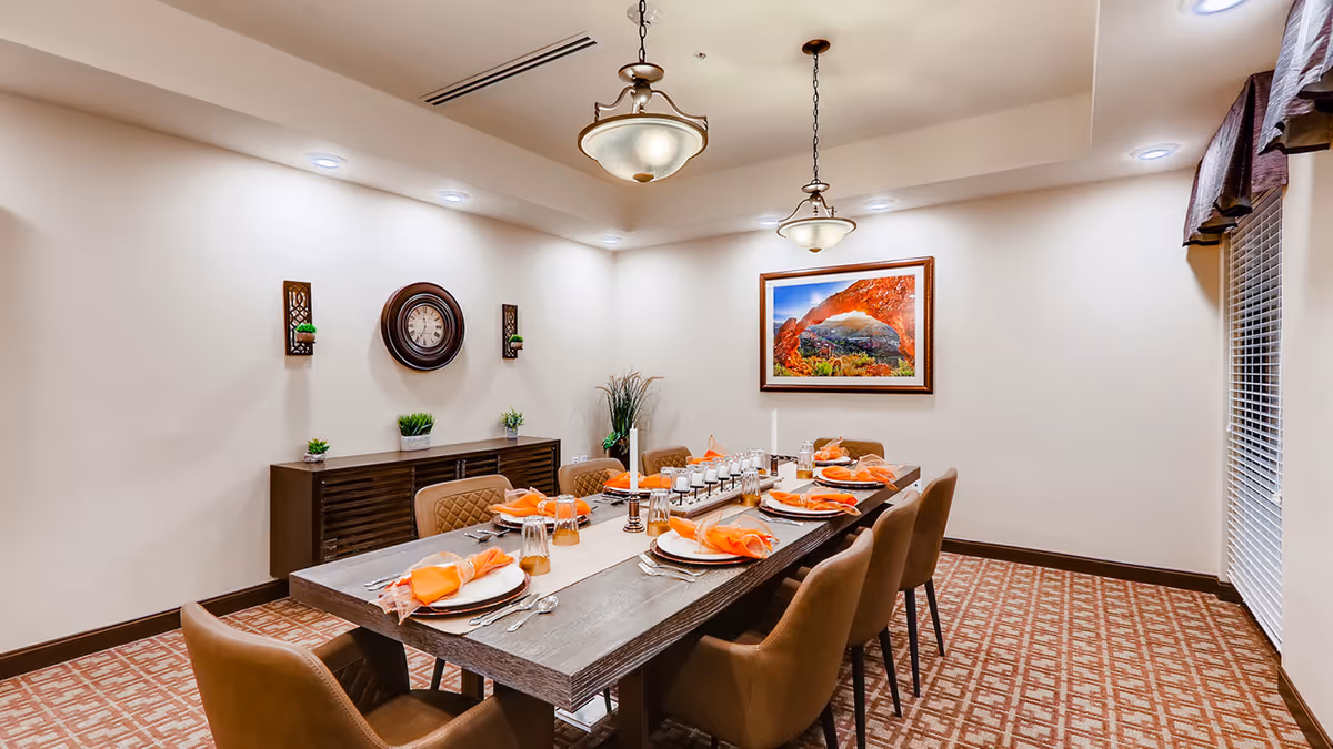 Long dining room table set for eight with orange napkins, pendant lights, framed art, and a sideboard.