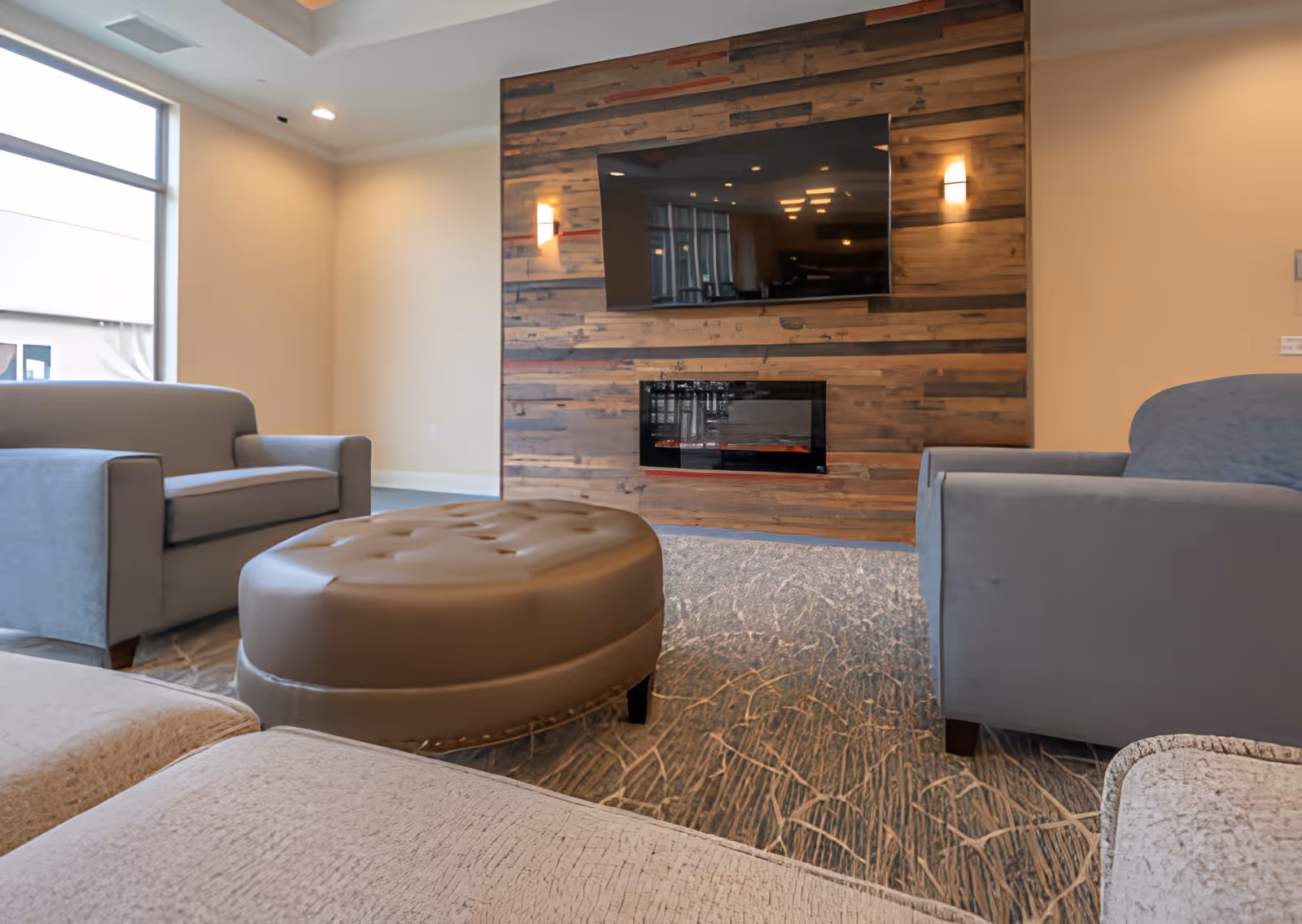 A cozy living room area with two gray armchairs and a large round brown ottoman in the center. The room features a wooden accent wall with a mounted flat-screen TV above a modern electric fireplace. There is a large window on the left side letting in natural light.