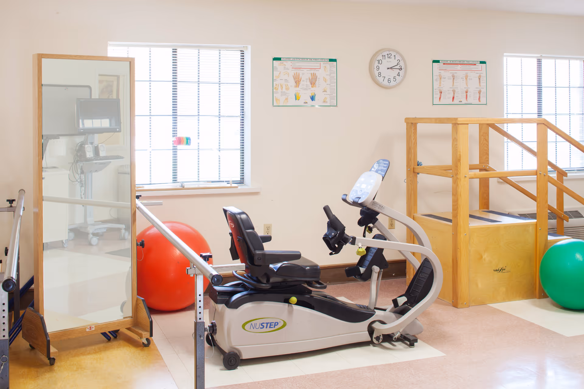A physical therapy room with a recumbent exercise bike, parallel bars, therapy stairs, exercise balls, and wall charts.