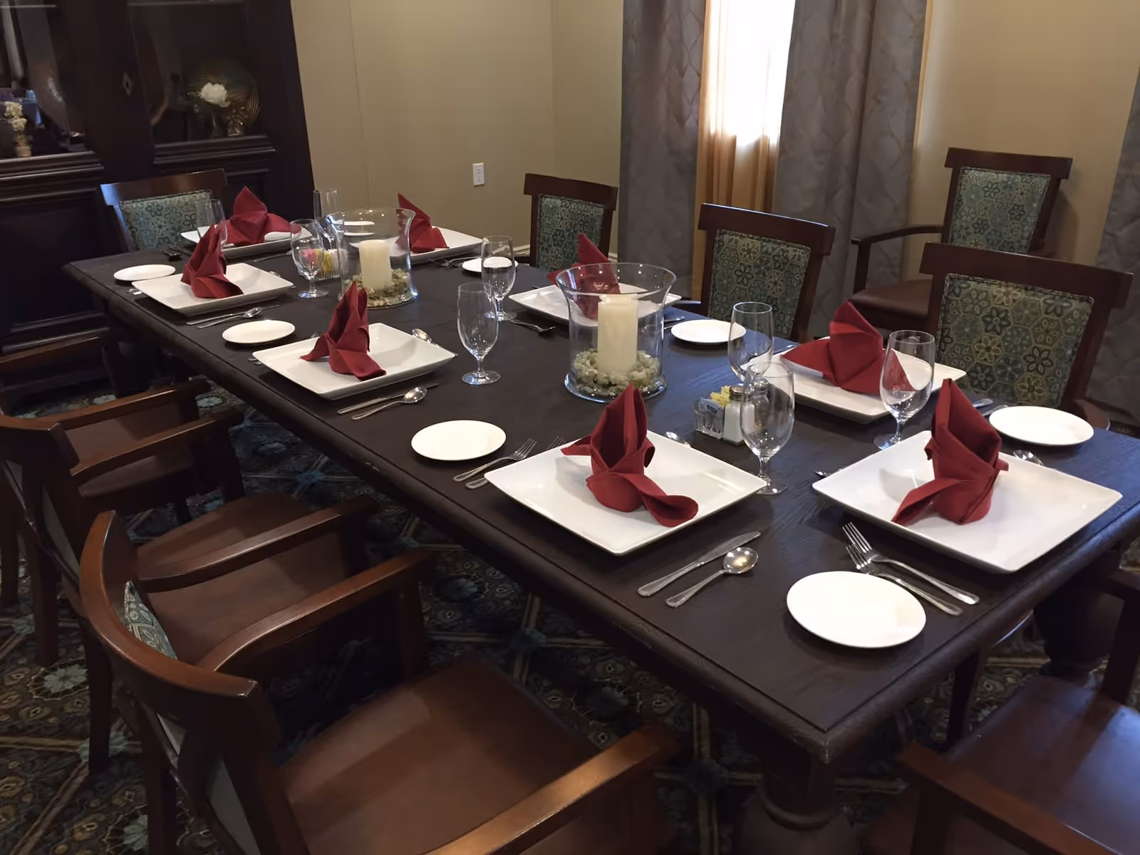 A dining room table set for eight people with white square plates, red folded napkins, water glasses, and silverware. The table has two glass candle holders with white candles and decorative stones inside. The chairs have wooden frames with patterned green cushions. Beige walls and curtains are visible in the background.