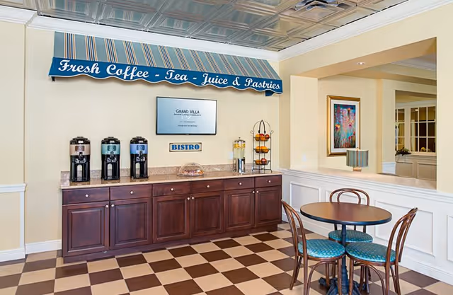 A small bistro area in a senior living facility with a countertop holding three coffee dispensers, a glass container with juice, and a tiered stand with pastries. Above the counter is a striped blue and white awning with the text 'Fresh Coffee - Tea - Juice & Pastries'. A digital screen and a small sign labeled 'BISTRO' are mounted on the wall. In front of the counter is a round table with four chairs with blue cushioned seats. The floor has a brown and beige checkered pattern, and the walls are painted light yellow with white wainscoting.