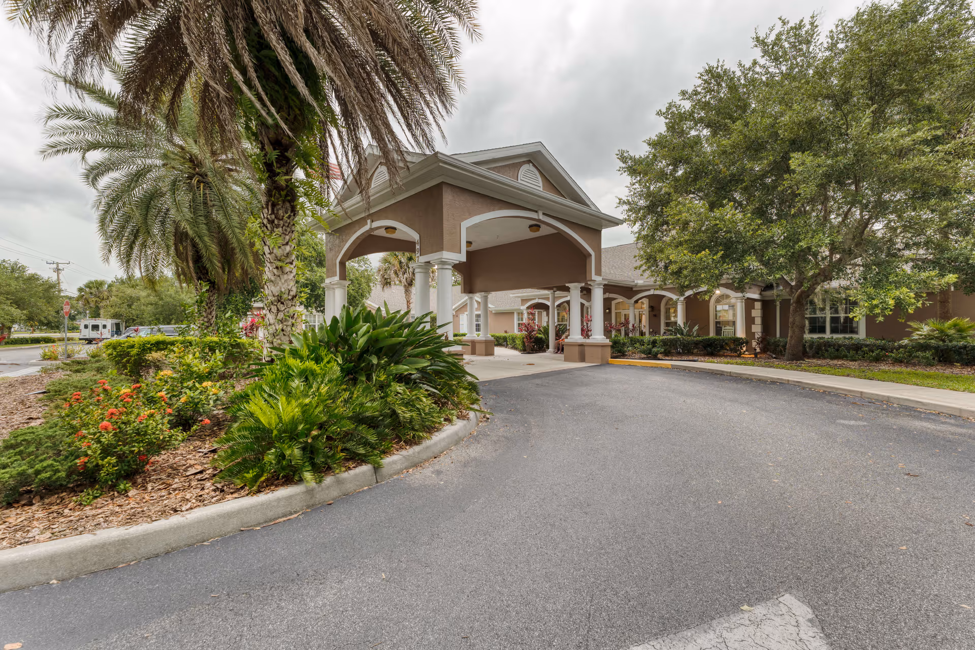 Front exterior view of Addington Place of Titusville showing a covered entrance with columns, surrounded by palm trees, bushes, and other greenery under a cloudy sky.