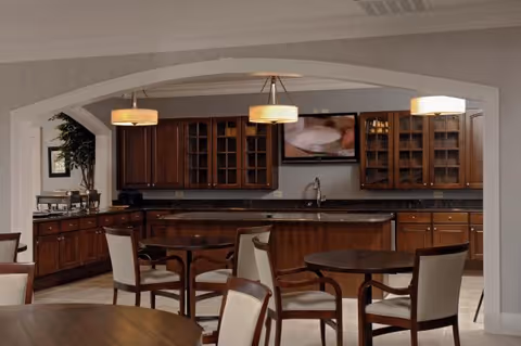 Communal dining area with round wooden tables and chairs facing a kitchenette with dark wood cabinets, pendant lights, and a wall-mounted TV.