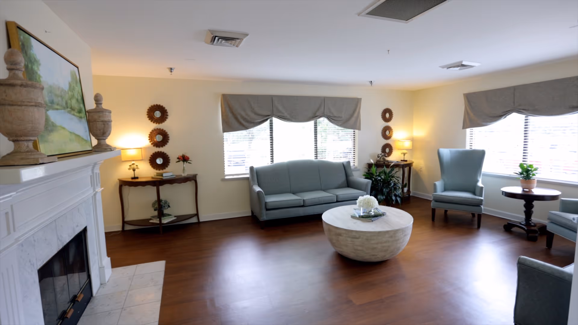 Bright assisted-living common room with a sofa and armchairs around a round coffee table, a fireplace on the left, and windows with valances.