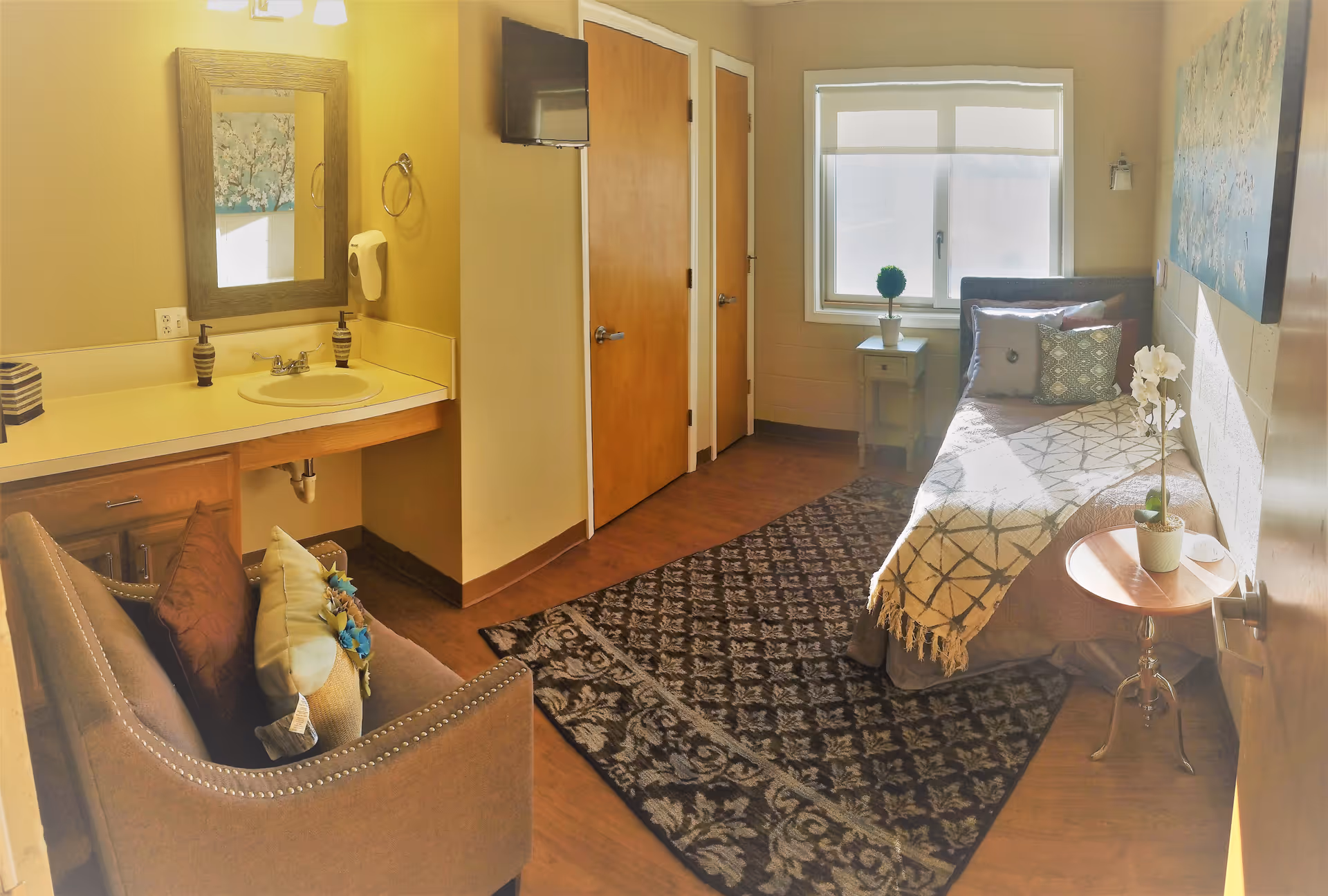 A cozy senior living room with a single bed covered with a patterned blanket and decorative pillows, a small round wooden side table with a potted plant, a window with blinds letting in natural light, a patterned area rug on the wooden floor, a brown armchair with cushions, and a sink with a mirror and soap dispenser on the left wall.
