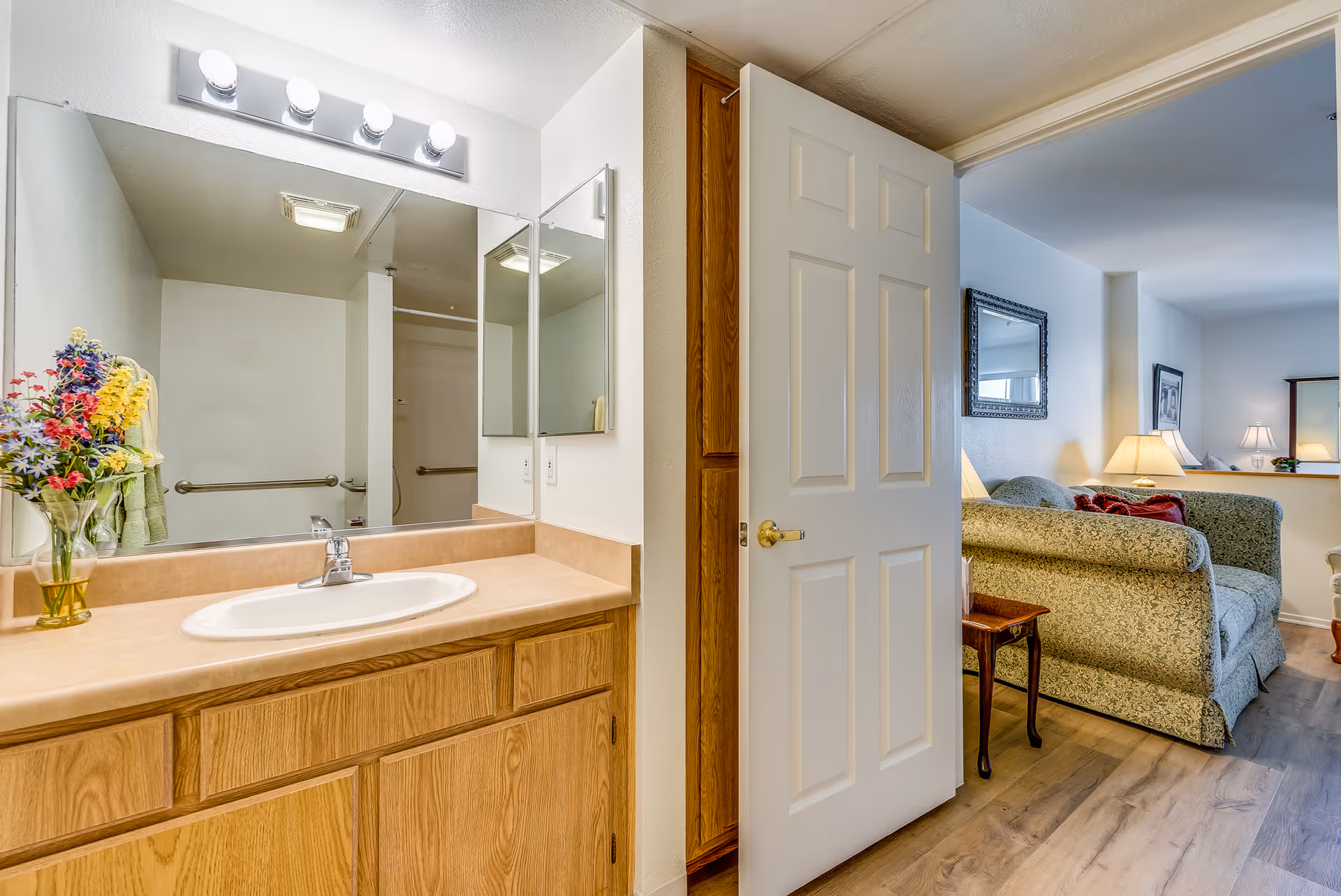 View of a bathroom vanity with a sink, wooden cabinets, and a large mirror with four lights above it. A vase with colorful flowers is placed on the countertop. The bathroom door is open, revealing a living room area with a patterned sofa, side table, lamps, and framed artwork on the walls.
