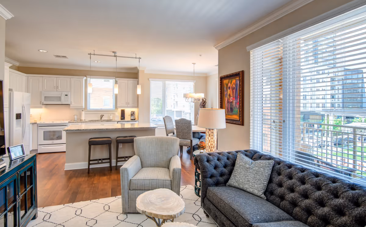 Bright and modern open-concept living space featuring a kitchen with white cabinets, a breakfast bar with two stools, a dining area with a table and chairs, and a living room area with a gray tufted sofa, an armchair, a small round coffee table, a floor lamp, and large windows with white blinds letting in natural light.