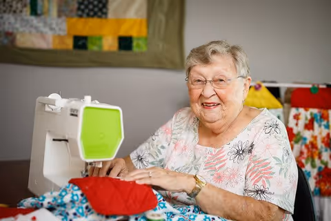 An elderly woman with glasses and a floral shirt smiling while using a sewing machine to work on a colorful quilt in a well-lit room with other quilts hanging in the background.
