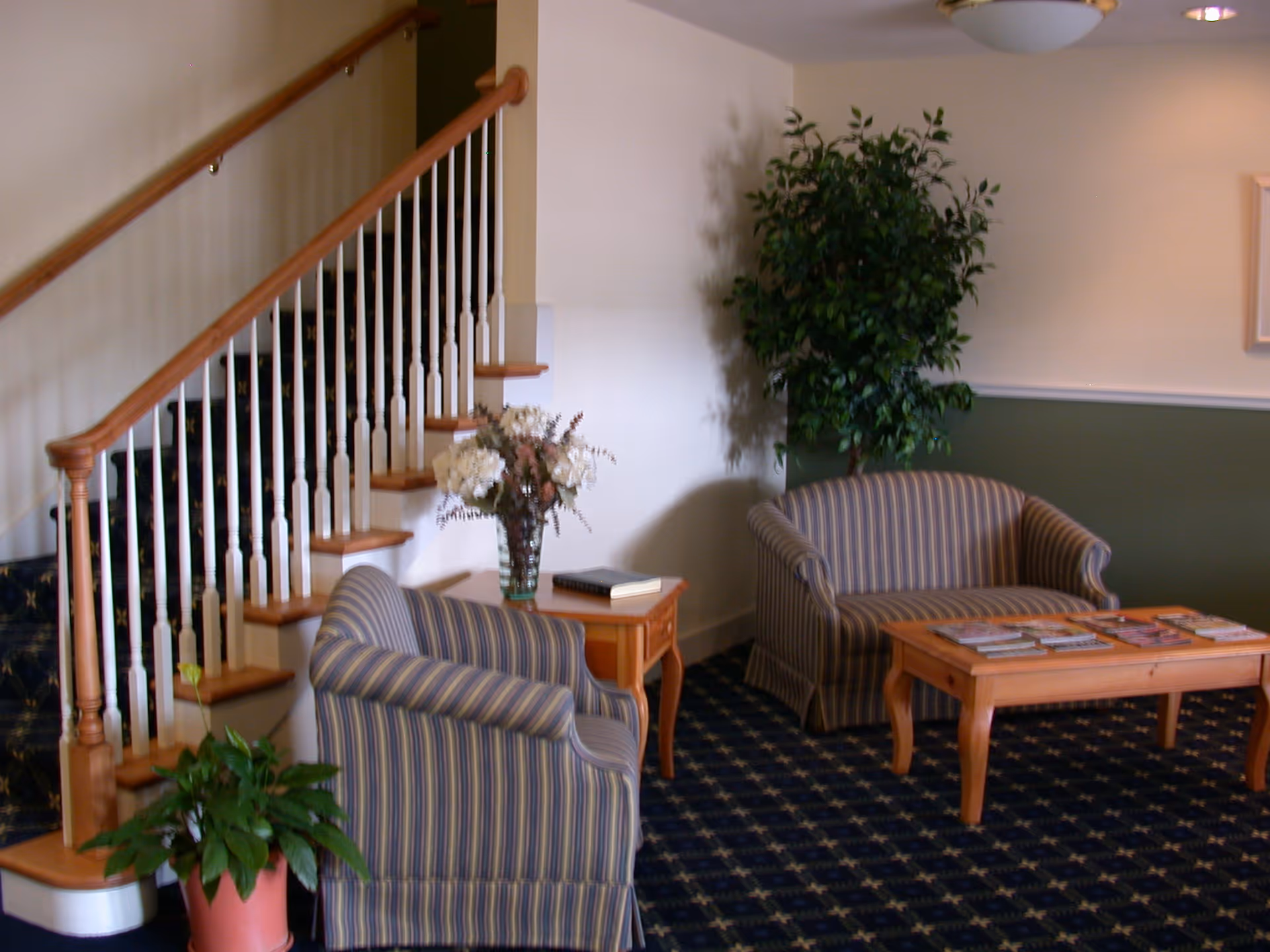 A cozy sitting area with striped armchairs and a loveseat, a wooden coffee table, potted plants, and a staircase in the background.