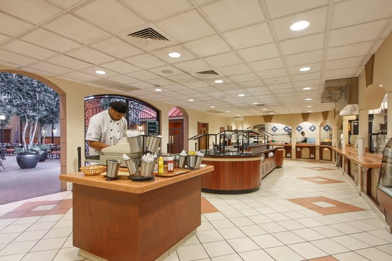 A spacious dining area with a buffet setup featuring multiple counters and food stations. A chef is standing behind one of the counters preparing or serving food. The area has tiled floors with a diamond pattern and a drop ceiling with recessed lighting. There are large arched openings leading to an outdoor patio with trees and seating visible outside.