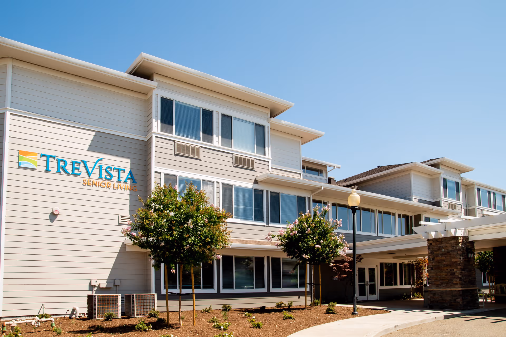Exterior view of TreVista Senior Living facility showing a multi-story building with large windows, a covered entrance supported by stone pillars, small trees and landscaping in front, under a clear blue sky.