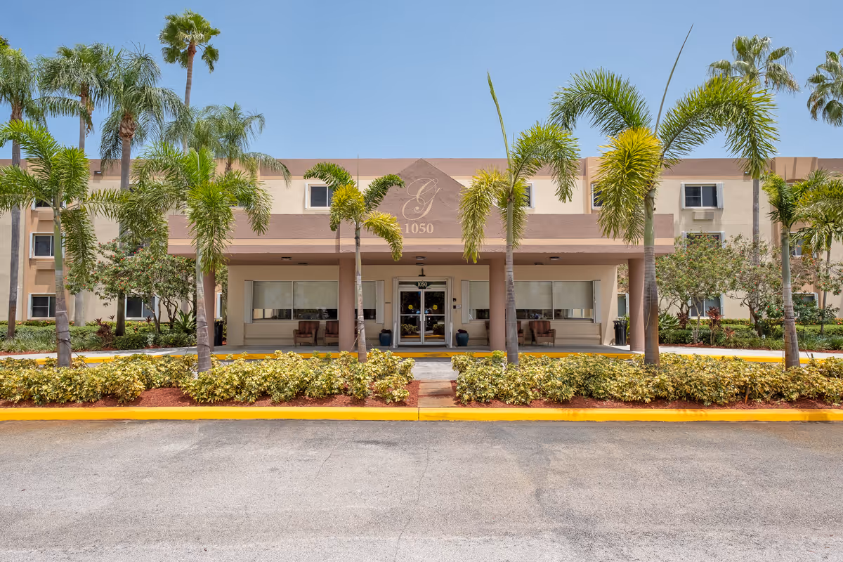 Front exterior view of Grand Villa of Deerfield Beach building with palm trees, shrubs, and a covered entrance under a clear blue sky.