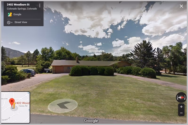 Street view of a single-story building with a sloped roof surrounded by green bushes and trees. There is a large grassy lawn in front of the building and a driveway with parked cars on the left side. The sky is partly cloudy.