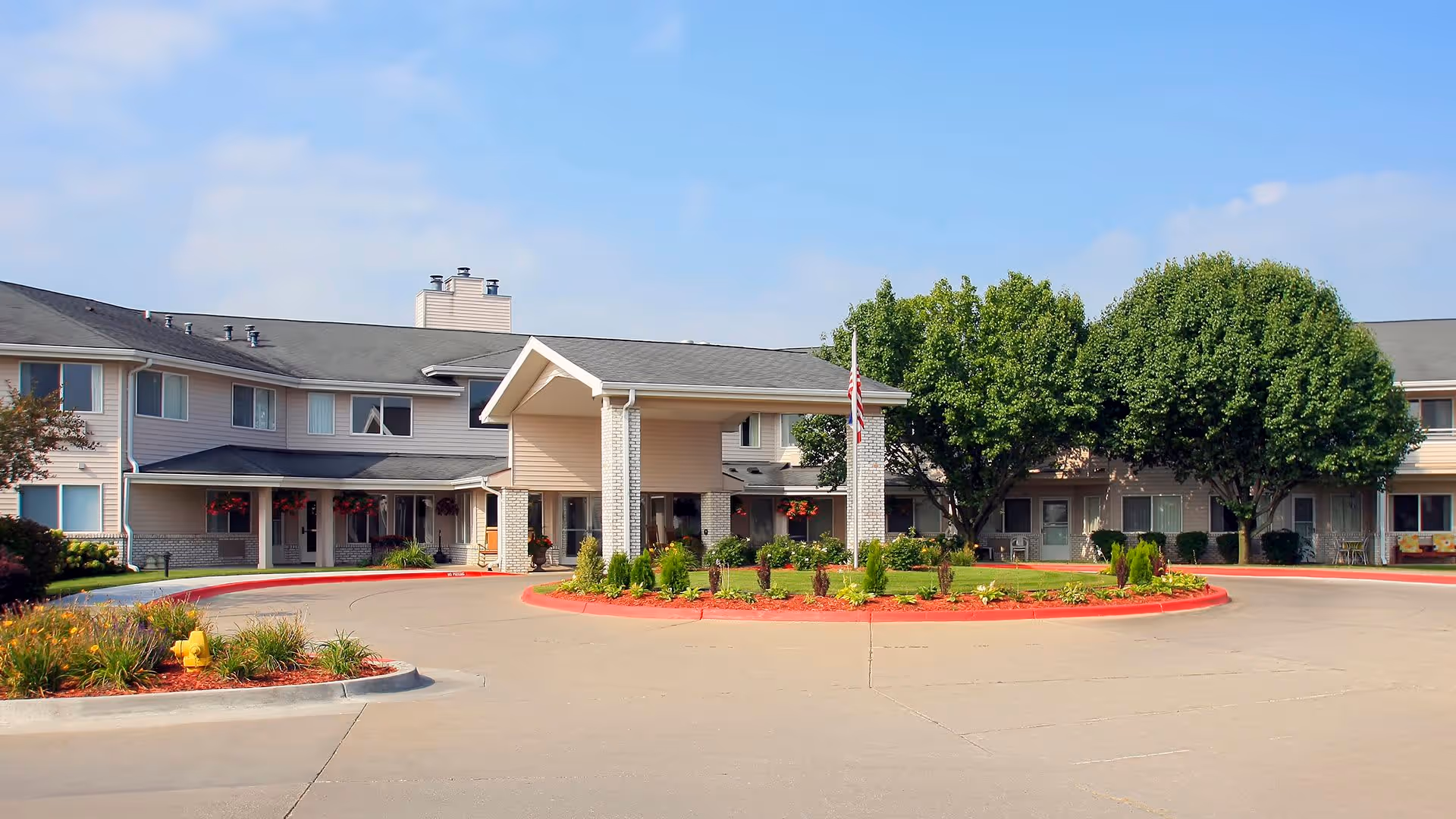 Front exterior view of Holiday Illahee Hills facility showing a two-story building with a covered entrance, a circular driveway, landscaped greenery, and trees under a partly cloudy sky.