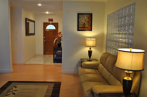 Interior view of a living room area with a beige leather sofa, two table lamps with beige shades, a framed floral painting on the wall, and a glass block window. In the background, there is a hallway leading to a wooden door with an exit sign above it, a mirror on the left wall, and a small table with a flower arrangement.