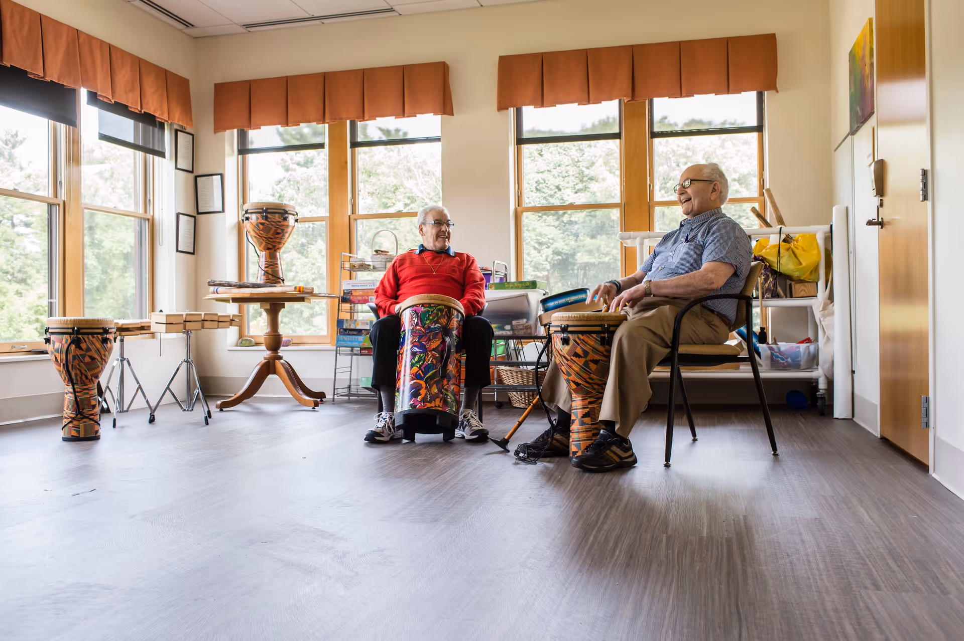 Two elderly men sitting in a bright room with large windows, playing colorful drums and smiling at each other. The room has wooden floors, orange valances on the windows, and various musical instruments and supplies around.