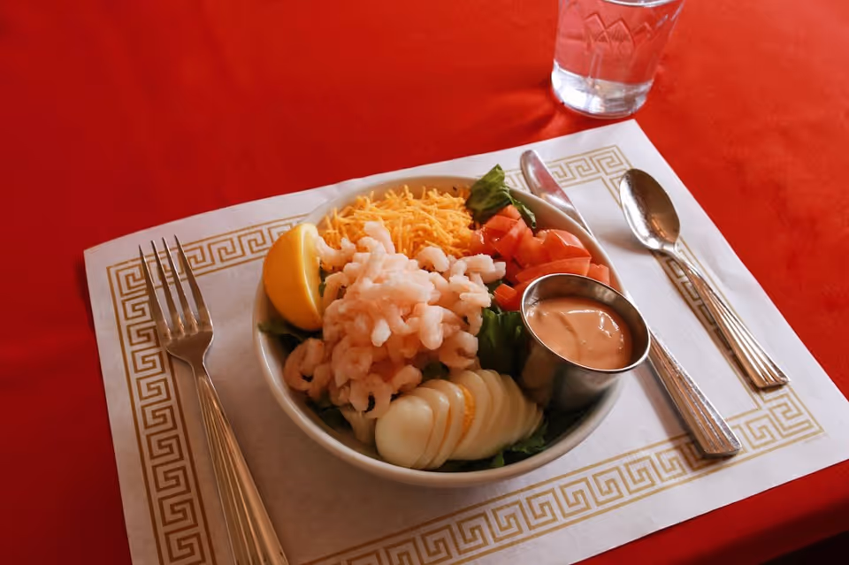 A bowl of shrimp salad with sliced egg, shredded cheese, diced tomatoes, a lemon wedge and a cup of dressing on a placemat with fork, spoon and a glass of water on a red tablecloth.