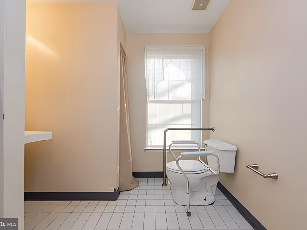 A bathroom with beige walls and white tiled floor. There is a white toilet equipped with safety rails on both sides. A window with sheer white curtains is located behind the toilet. A metal grab bar is mounted on the right wall. The room is well-lit with natural light coming through the window.