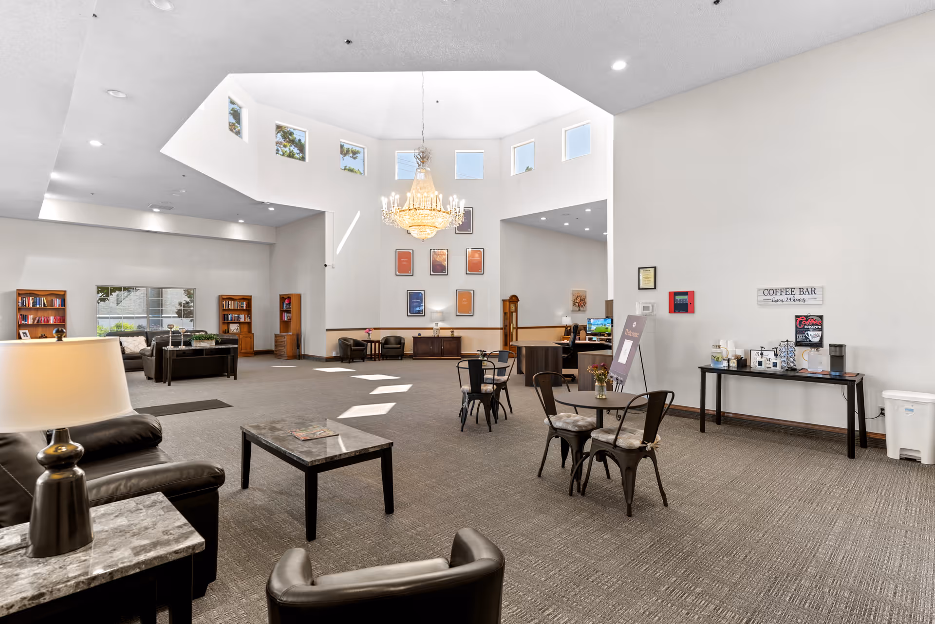 Spacious senior living facility common area with high ceilings and multiple windows near the ceiling allowing natural light. The room features a chandelier, several seating areas with chairs and tables, bookshelves, a coffee bar station, and a reception desk in the background.
