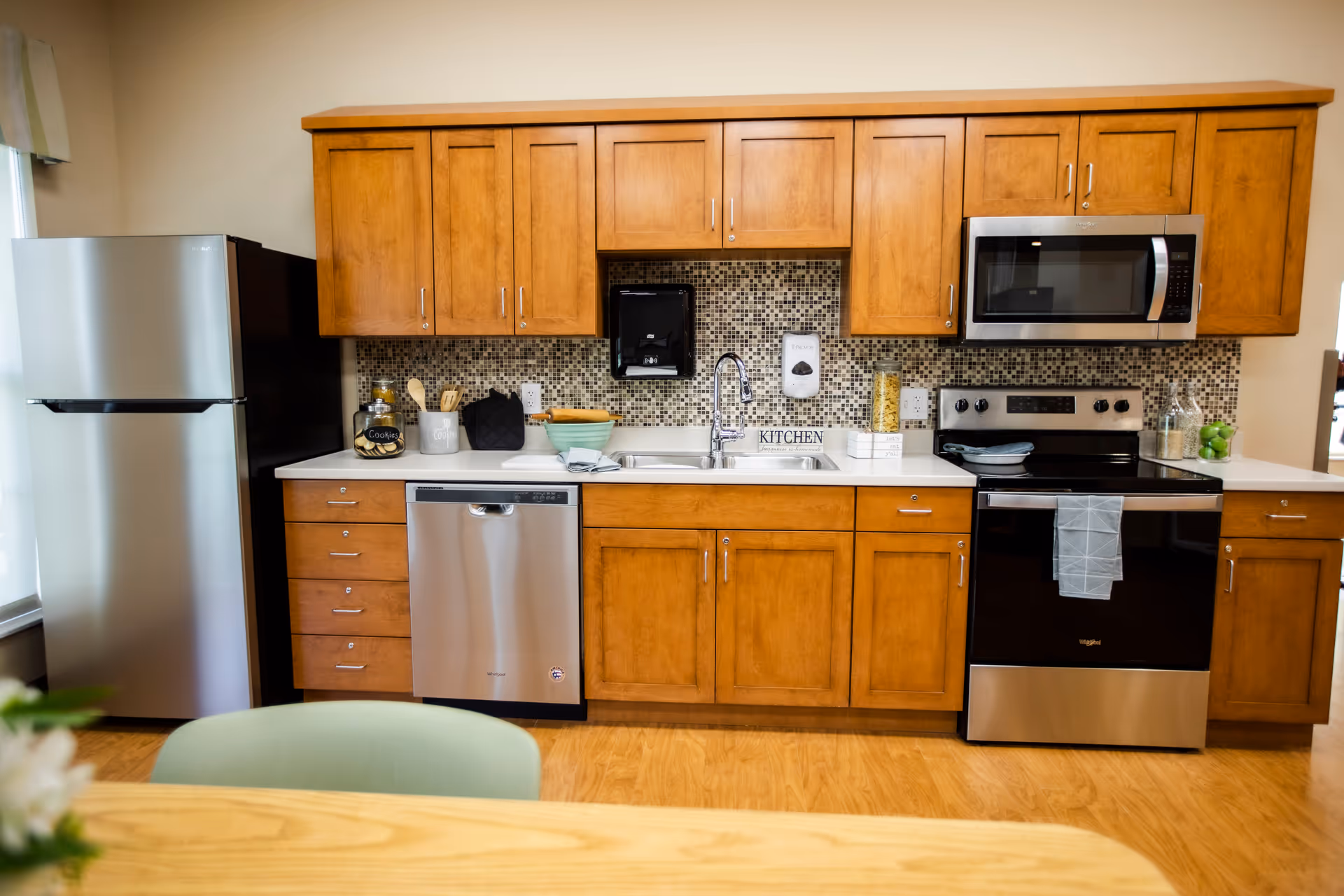 A modern kitchen with wooden cabinets, a stainless steel refrigerator, dishwasher, stove, and microwave. The backsplash features small square tiles in neutral tones. There is a sink with a chrome faucet, a cookie jar, kitchen utensils, and decorative items on the countertop. A wooden table and a green chair are partially visible in the foreground.