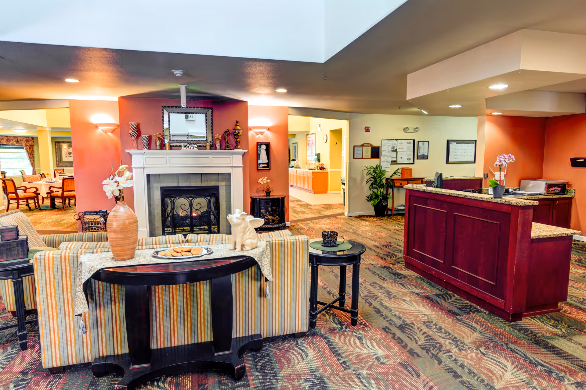 Interior common area with a reception desk, seating with a striped sofa facing a fireplace, and a dining area visible in the background.