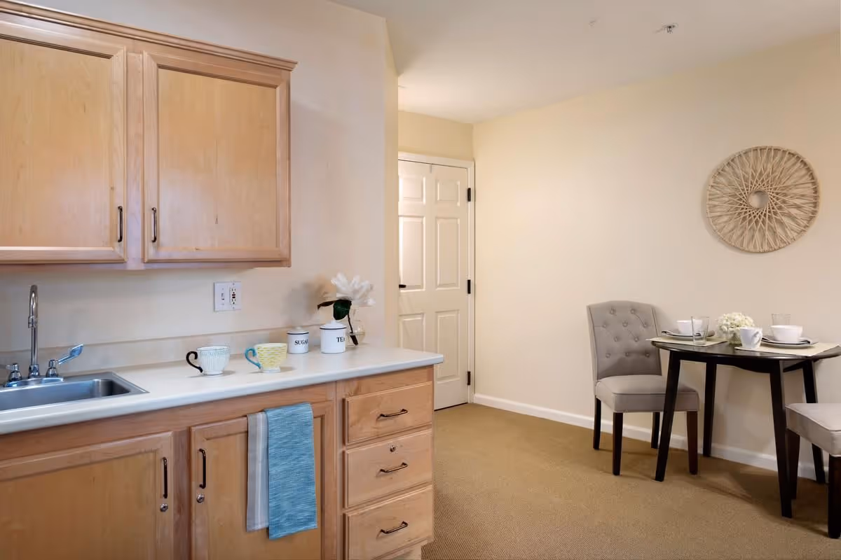 A small kitchen area with light wood cabinets, a stainless steel sink, and a countertop holding three cups and two labeled containers for sugar and tea. A blue towel hangs from a drawer handle. Adjacent to the kitchen is a small dining area with a dark wooden table set for two, featuring white dishes and a small flower arrangement. The walls are light beige, and there is a decorative circular wall hanging above the dining table.