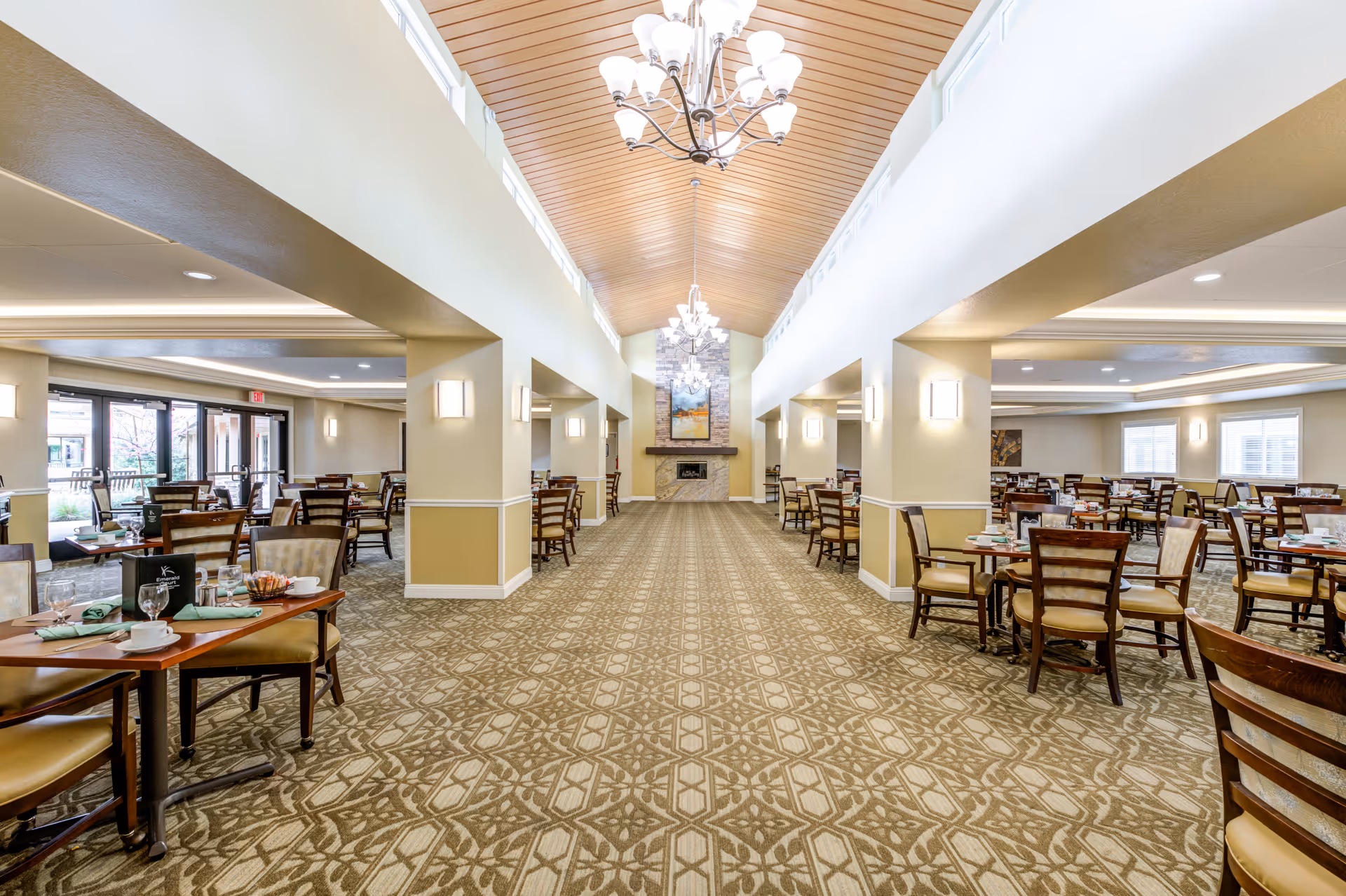 Spacious dining room with multiple wooden tables and chairs arranged neatly. The room features a patterned carpet, beige walls with white trim, and a high wooden ceiling with chandeliers. Large windows and glass doors allow natural light to enter, and a fireplace with artwork above it is visible at the far end.