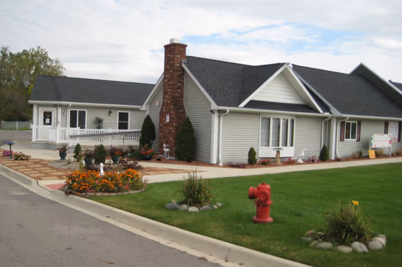 Single-story light-gray building with a brick chimney, porch, landscaped flowerbeds and a red fire hydrant on the lawn in front.