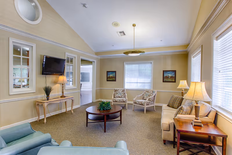 A cozy living room with beige walls and carpeted floor, featuring a beige sofa with patterned cushions, two armchairs with floral upholstery, a round wooden coffee table with a plant centerpiece, a wooden side table with a lamp, a wall-mounted flat-screen TV above a small wooden console table with a lamp and plant, and three windows with white blinds letting in natural light.