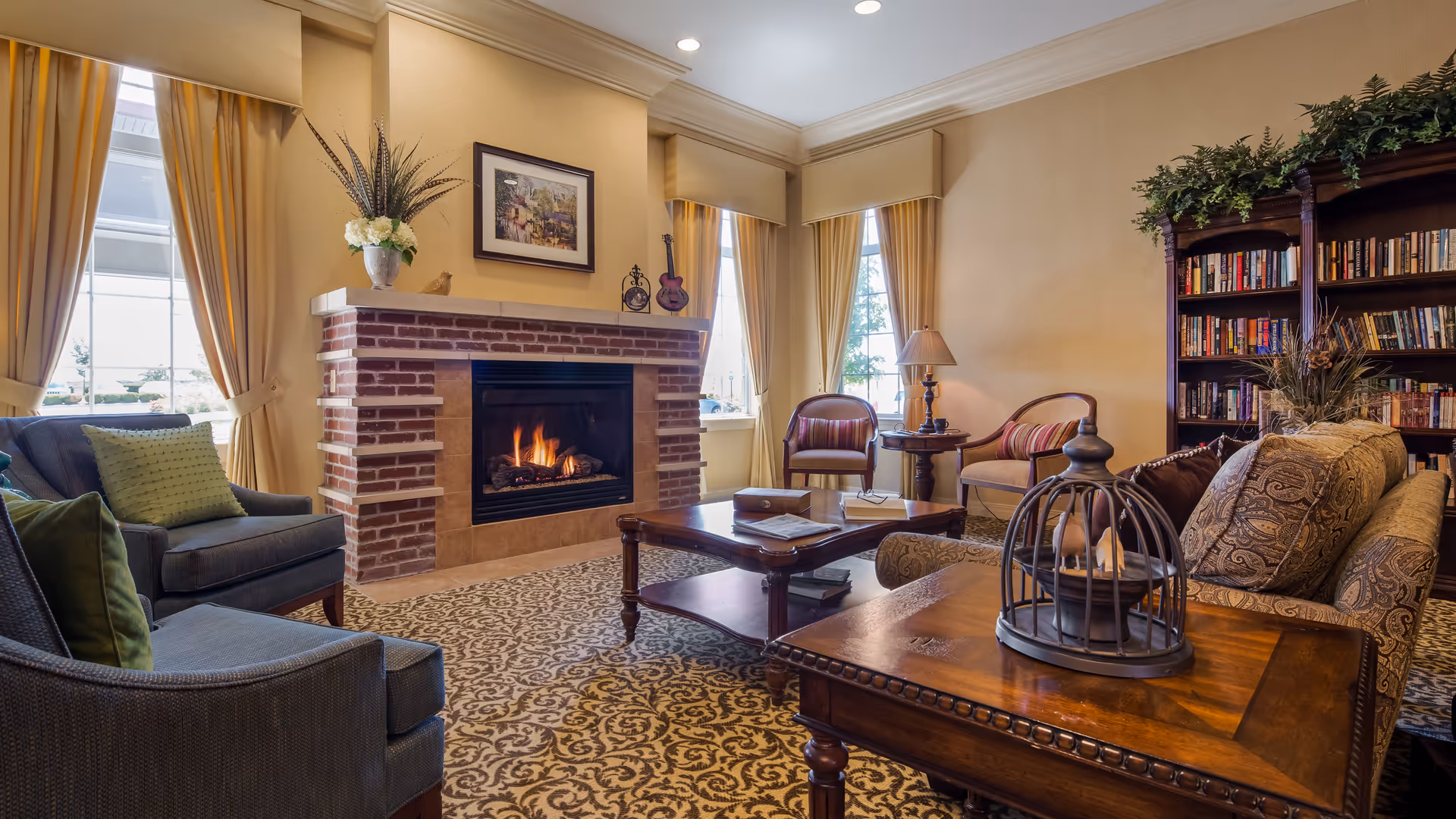 A cozy living room with a lit brick fireplace, two large windows with beige curtains, comfortable armchairs and a sofa with patterned cushions, a wooden coffee table, a side table with a lamp, and a bookshelf filled with books. The room has warm lighting and a patterned carpet.