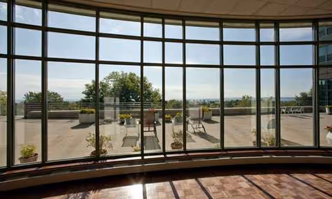 Curved glass wall opens to a sunlit outdoor patio with tables, chairs, and planters.