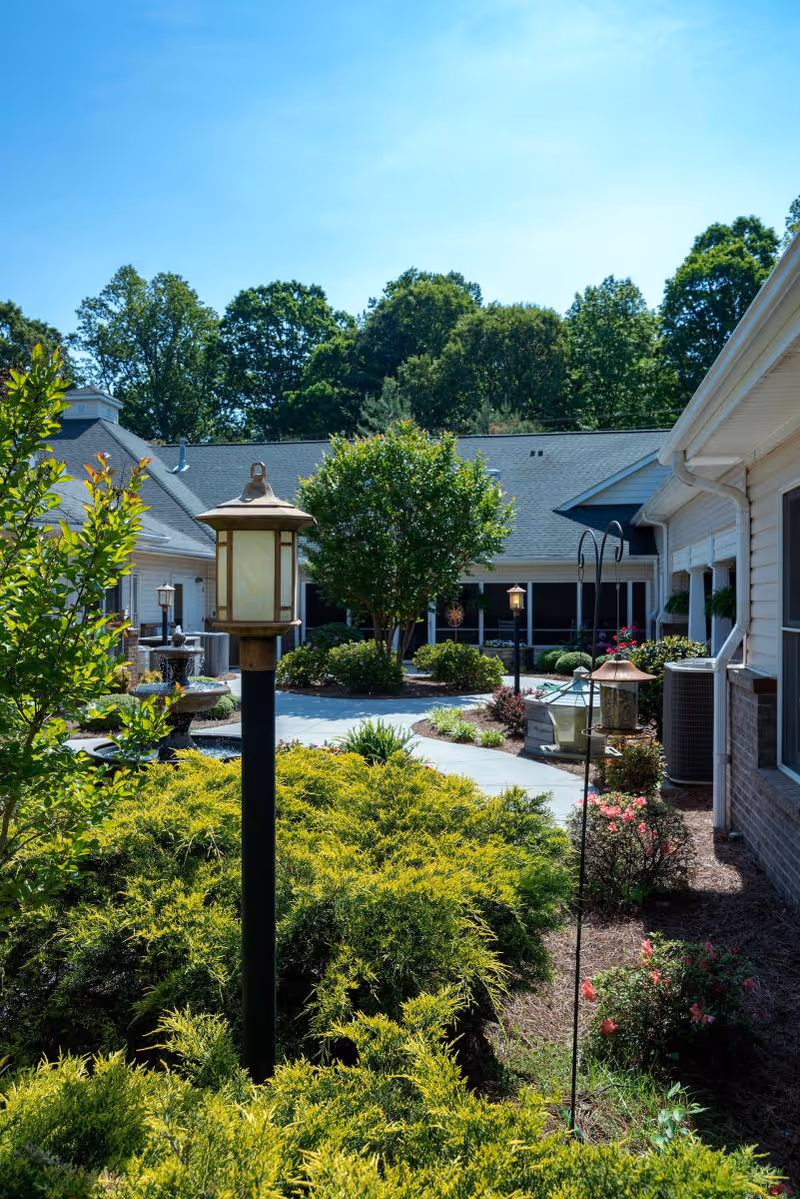 Outdoor courtyard area at Brookstone of Clemmons featuring a paved walkway, green shrubs, trees, decorative lamp posts, and a water fountain, surrounded by building structures under a clear blue sky.