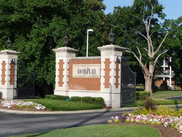 Entrance gate to AdamsPlace facility with brick and stone pillars, black wrought iron gates, surrounded by green trees and landscaped flower beds with colorful flowers.