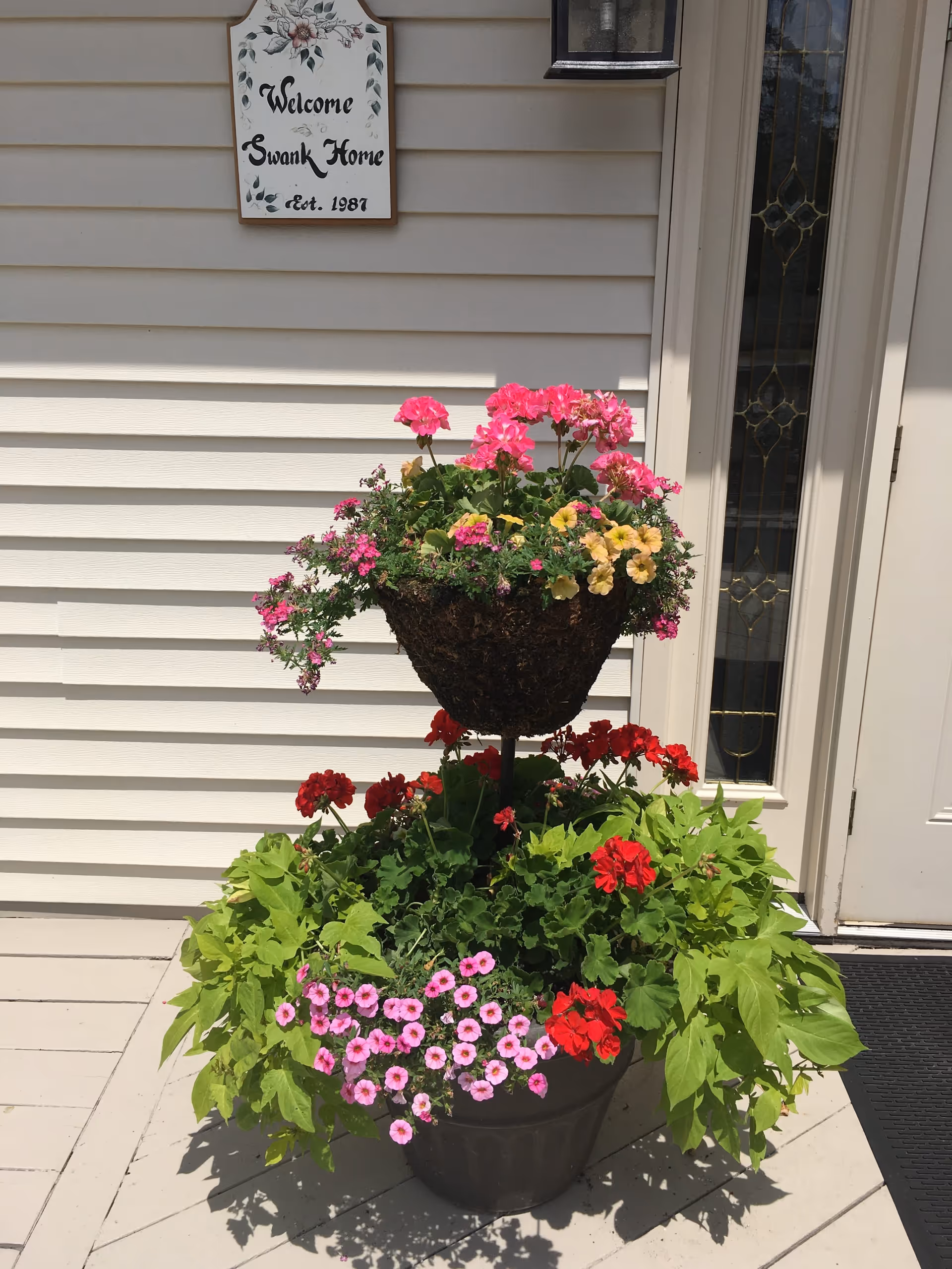 A decorative flower arrangement with pink, red, and yellow flowers in a two-tiered planter placed on a light-colored wooden porch. Behind the planter is a beige siding wall with a sign that reads 'Welcome Swank Home Est. 1987' and a white door with a decorative glass panel.