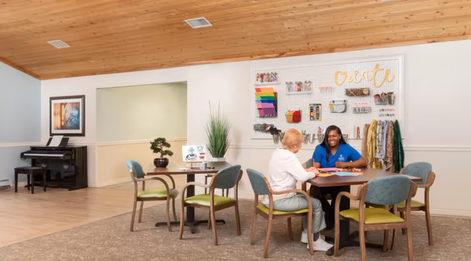 A senior woman and a caregiver sitting at a table in a bright, welcoming activity room. They are engaged in a creative craft activity with colorful paper. The room features a wooden ceiling, a pegboard wall organizer with craft supplies, a piano in the corner, and several chairs around tables.
