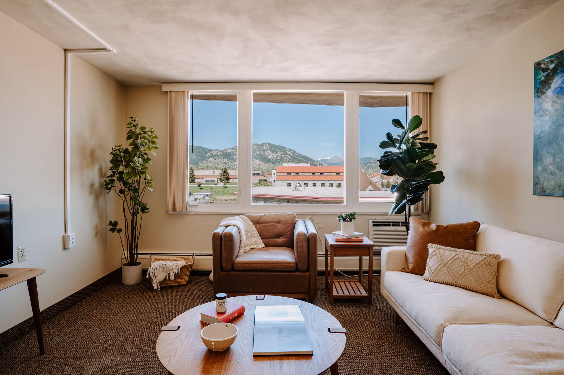 A cozy living room with a large window showing a mountain view. The room features a brown leather armchair, a beige sofa with decorative pillows, a wooden coffee table with a bowl, a book, and a cup on it. There are two potted plants, one tall in the corner and one on a side table next to the armchair. A colorful abstract painting hangs on the wall.