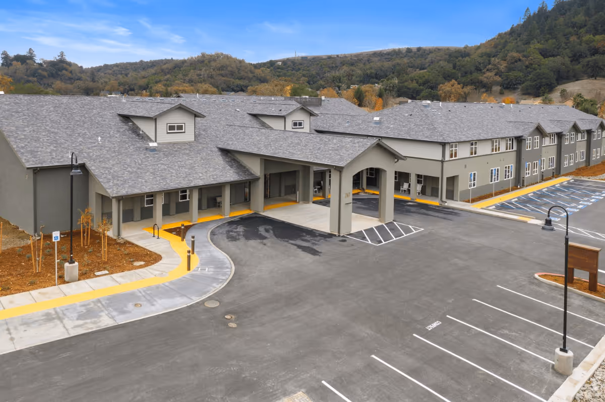 Exterior view of Vine Ridge at Cloverdale facility showing a large, modern building with a covered entrance and an adjacent parking lot. The building has a gray roof and beige walls, surrounded by landscaped areas and hills in the background under a partly cloudy sky.