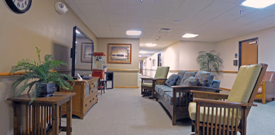 A hallway in a retirement community with a seating area including a patterned sofa and two wooden armchairs with cushions. There is a wooden side table with a potted plant, a TV on a wooden stand, a popcorn machine, framed artwork on the wall, and a clock. The hallway is carpeted and has ceiling lights and doors along the walls.