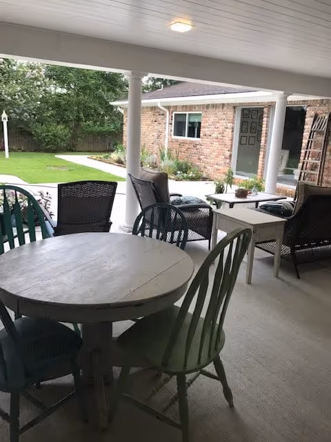 Covered patio with a round wooden table and chairs, wicker lounge seating, and a view of a brick building and lawn.